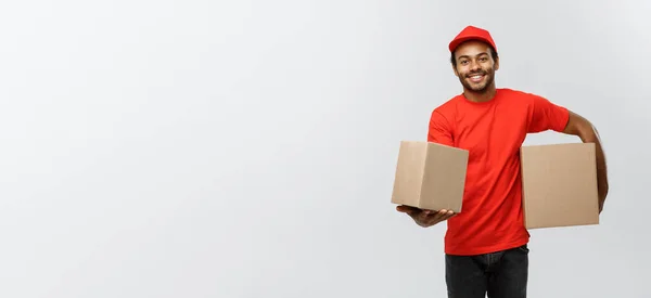 Conceito de entrega - Retrato de homem de entrega afro-americano feliz em pano vermelho segurando um pacote de caixa. Isolado no estúdio Grey Background. Espaço de cópia . — Fotografia de Stock