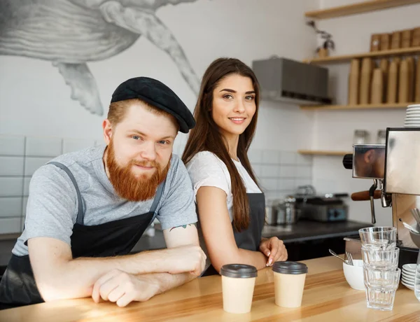 Coffee Business Concept - Positive young bearded man and beautiful attractive lady barista couple in apron while standing at bar Couter ready to give Coffee Service at the modern coffee shop — Stockfoto