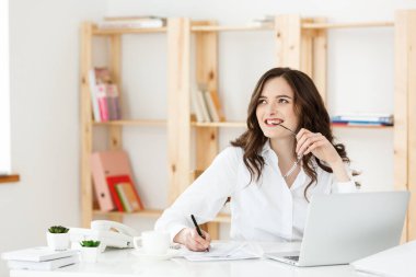 Modern bir ofis masasında dizüstü bilgisayarla çalışan ve bir şeyler düşünen çekici genç bir kadın..