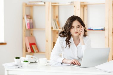 Modern bir ofis masasında dizüstü bilgisayarla çalışan ve bir şeyler düşünen çekici genç bir kadın..