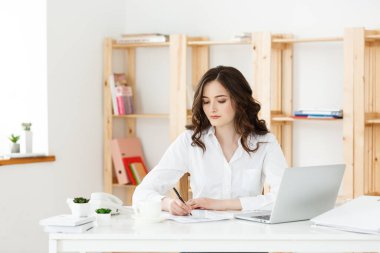 Modern bir ofis masasında dizüstü bilgisayarla çalışan ve bir şeyler düşünen çekici genç bir kadın..