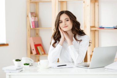 Modern bir ofis masasında dizüstü bilgisayarla çalışan ve bir şeyler düşünen çekici genç bir kadın..