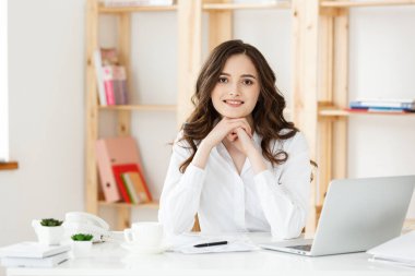 Modern bir ofis masasında dizüstü bilgisayarla çalışan ve bir şeyler düşünen çekici genç bir kadın..