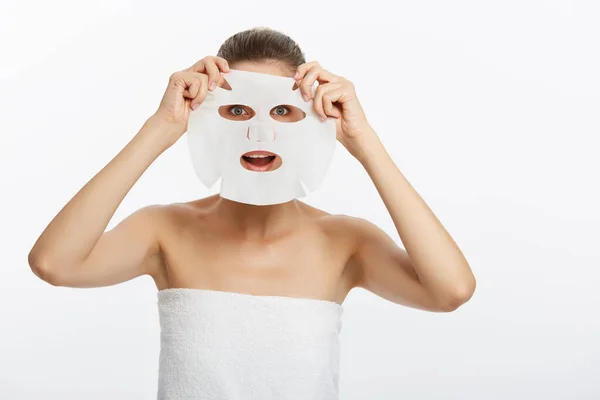 Mladá dívka s obličejovou maskou dívá na kameru na bílém pozadí. Kosmetická procedura. Kosmetické lázně a kosmetologie — Stock fotografie