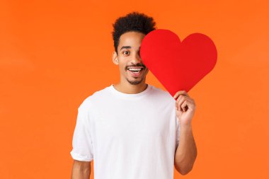 Sevgililer günü, ilişki ve takdir konsepti. Çekici, romantik erkek arkadaş, Afro-Amerikan adam, kalp kartını yüzüne yakın tutuyor ve mutlu bir şekilde gülümsüyor, gerçek aşkı arıyor, turuncu arka plan.