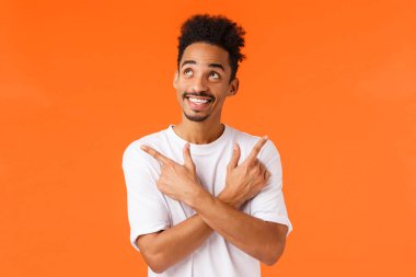 Hevesli, heyecanlı, bıyıklı Afro-Amerikan erkek, rüya gibi ve tasasız, mutlu bir şekilde gülümseyen, sağı solu işaret eden, seçim yapan, ne alacağına karar veren, turuncu arka planlı..