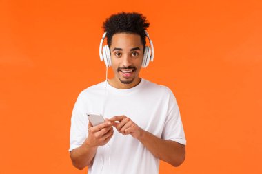 Heyecanlı, mutlu, karizmatik, Afro saçlı, bıyıklı, Afro saçlı Amerikalı hippi adam elinde akıllı telefon, step aleti seçmeli şarkı, büyük kulaklıklarla müzik dinliyor, turuncu arka plan..