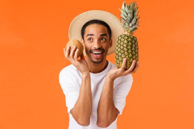 Neşeli ve mutlu, gülümseyen Afro-Amerikalı turist şapkalı, beyaz tişörtlü, hindistan cevizi ve ananas tutan, heyecan ve neşe gösteren, yaz tatilinde dinlenen, yurt dışına seyahat eden.