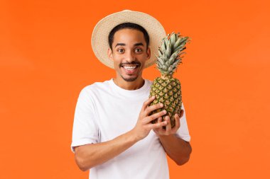 Tatilin tadını çıkarma zamanı. Mutlu gülümseyen, Afro-Amerikan erkek turist, ananas tutan ve gülümseyen erkek arkadaş, yurtdışı gezisi sırasında yerel meyveleri deniyor, lüks oteller, turuncu arka plan..