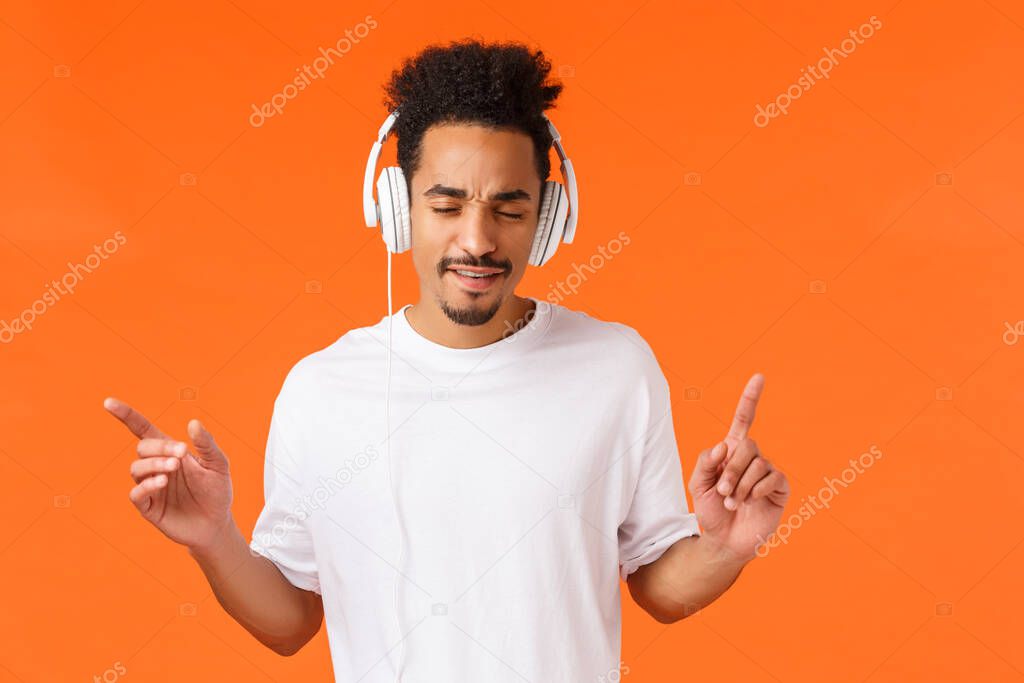 Man enjoying awesome beats. Attractive modern hipster african-american guy with afro haircut, moustache, close eyes drumming with fingers and listen music in headphones, orange background.