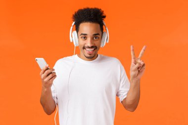 Barışçıl ve dost canlısı, beyaz tişörtlü mutlu Afro-Amerikan adam, müzik kulaklıkları, barış işareti ve gülümseme gösteren, akıllı telefon tutan, arkadaşlarının şarkı listesini dinleyen, yeni çıkan.