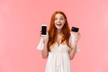 Çekici, keyifli ve büyüleyici mutlu kızıl saçlı kız mutlu ve mutlu, elinde kredi kartıyla, akıllı telefon ekranını göstererek, harika bir alışveriş yaptı, arkadaşına böbürlenerek, pembe arka planda.