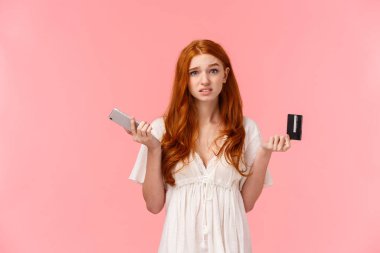 Kararsız ve kararsız Avrupalı tatlı kızıl saçlı kadın omuz silkiyor, kafası karışmış ve işe yaramaz kameraya bakıyor, elinde akıllı telefon ve kredi kartıyla omuz silkiyor, hangi sırayla çevrimiçi bilmiyorum..