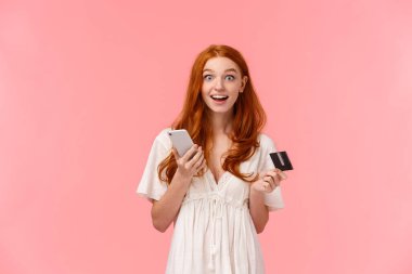 Etkilenmiş ve heyecanlı, beyaz elbiseli mutlu kızıl saçlı kız, ne kadar kolay satın aldığına şaşırmış, elinde akıllı telefon ve kredi kartı, gülümsüyor ve merak uyandıran kamera, pembe arka plan..