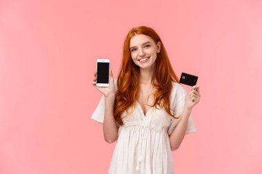 Mobil ekranda uygulamayı tanıtan kız. Neşeli, güzel kızıl saçlı beyaz kadın kredi kartı tutuyor ve akıllı telefon ekranını gülümseyerek gösteriyor. Uygulama ya da internet mağazası tanıtıyor..