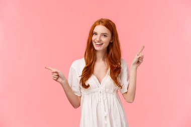 Happy alluring redhead female in white dress, helping out with fashion advices, pointing fingers left and right company banners, advertisements, smiling friendly and confident, showing what pick.
