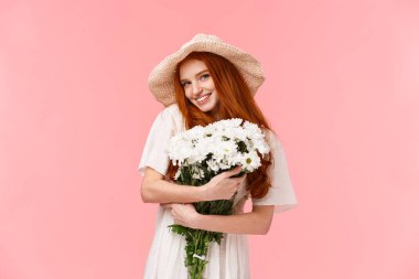 Güzel ve cezbedici romantik sevimli, saman şapkalı aptal kızıl kız, güzel beyaz çiçekleri kucaklıyor, buket hediyesini alıyor ve gülümsüyor kamera küstahça, durgun pembe arka plan.