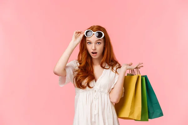 多么大胆的举动 惊慌失措 说不出话来 惊讶的红头发姑娘摘下眼镜 看到令人印象深刻的东西 放下下巴 背着购物袋 站在粉色的背景上 — 图库照片