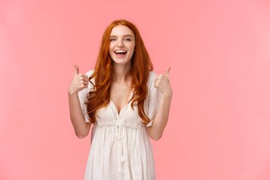 Belden yukarı, neşeli, neşeli, beyaz elbiseli kızıl saçlı kadın, başparmağını kaldırmış ve gülümsüyor, ürün gibi, tavsiye et, olumlu görüş bildir, pembe arka planda dur.