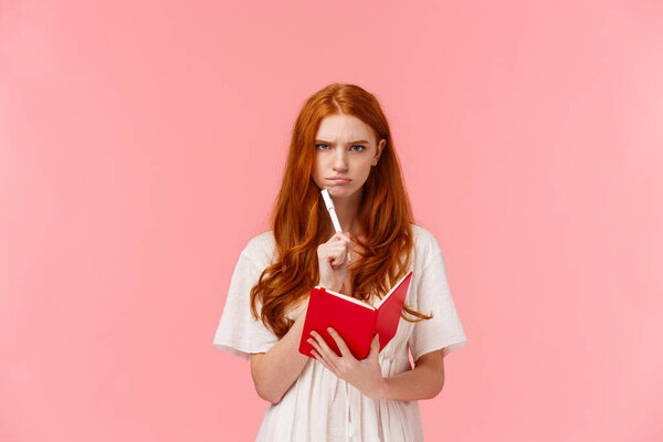 Serious-looking grumpy redhead woman frowning as thinking, pouting troubled, solving serious riddle, holding red notebook and touching chin with pen, pondering over homework, pink background.