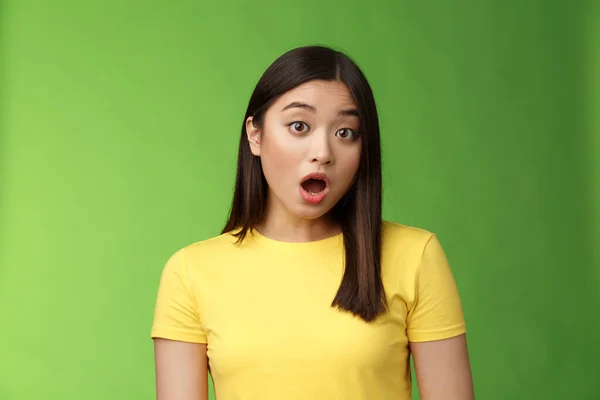 Close-up shocked amazed asian brunette female astonished hear stunning gossip, drop jaw stare camera speechless stand stupor green background, surprised what happening, wear yellow t-shirt.