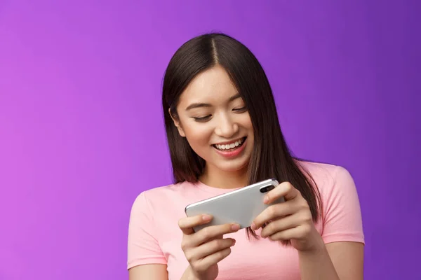 Close-up joyful attractive asian woman brunette having fun spend time playing smartphone game, laughing smiling eager win race, hold phone horizontal beating score, purple background.