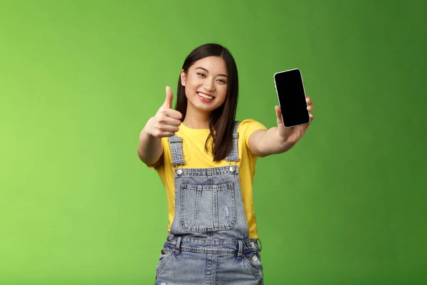 快乐满足的可爱的亚洲黑发女孩喜欢令人敬畏的智能手机游戏 展示电话显示 像手势一样竖起大拇指 面带微笑 介绍令人敬畏的在线应用 绿色背景 — 图库照片