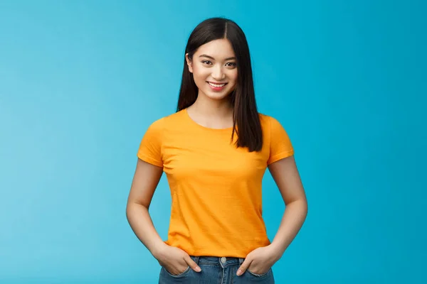 自信を持って積極的な笑顔アジアの女性は青の背景ルックカメラスタンド 手のジーンズのポケットを保持する自己表現安心した雰囲気 肯定的な結果のスキンケア手順を見て楽しむ 黄色のTシャツを着用 — ストック写真