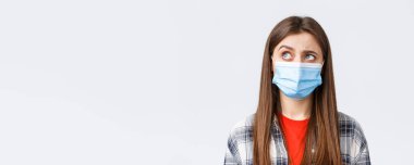 Coronavirus salgını, karantina, sosyal mesafeler ve duygu kavramı. Tıbbi maskeli, meraklı ve tereddütlü bir genç kadın sol üst köşeye bakıyor isteksiz, beyaz arka plan.
