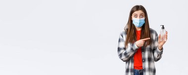 Coronavirus salgını, karantina, sosyal mesafeler ve duygu kavramı. Tıbbi maskeli, el dezenfektanını işaret eden heyecanlı ve şaşkın genç kadın,.