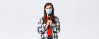 Coronavirus salgını, karantina, sosyal mesafeler ve duygu kavramı. Tıbbi maskeli kafası karışmış bir kadın. Elinde mikrofon, performans, beyaz arka plan..