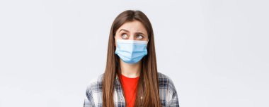 Coronavirus salgını, karantina, sosyal mesafeler ve duygu kavramı. Tıbbi maskeli, meraklı ve tereddütlü bir genç kadın sol üst köşeye bakıyor isteksiz, beyaz arka plan.