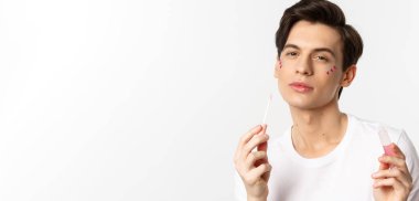 Yüzünde parıltı olan, dudak parlatıcısı süren, kameraya kendinden emin bir şekilde bakan, beyaz arka planda duran güzel, tuhaf bir insanın yakın çekimi..