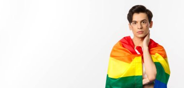 Yüzünde sim olan güzel bir gay adamın belden yukarı pozu, kendini gökkuşağı lgbt bayrağıyla sar ve yüzüne nazikçe dokun, kameraya bak, beyaz arkaplan.
