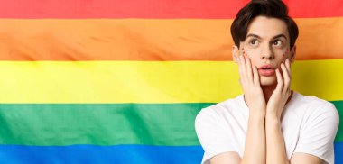 Yüzünde parıltı makyajı olan, sol üst köşeye bakan, heyecanlı, lgbtq gökkuşağı bayrağına karşı duran şehvetli ve güzel bir gay portresi..