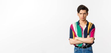 Gücenmiş ve kızgın genç eşcinsel adam somurtuyor, kollarını kavuşturuyor ve kameraya yargılayıcı bakıyor, beyaz arka planda duruyor..