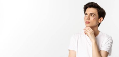 İnsanlar, lgbtq topluluğu ve yaşam tarzı konsepti. Güzel çift cinsiyetli, yüzünde sim olan adam, sol üst köşedeki şehvetli adama bakıyor, beyaz arka planda duruyor..