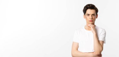 Beyaz tişörtlü, yüzünde sim olan ve kameraya ciddi ciddi bakan beyaz arka planda duran çekici bir gay resmi..