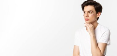 Millet, lgbtq ve güzellik konsepti. Güzel çift cinsiyetli adam, gözlerinin altında pırıltı, sol üst köşeye bakıyor, beyaz arka plana karşı poz veriyor..