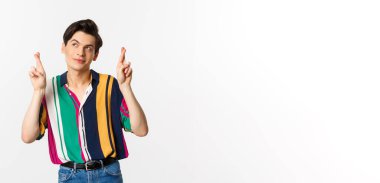 Hayalperest genç adam parmaklarını çaprazlayarak dilek diliyor, gülümsüyor ve sol üst köşeye bakıyor düşünceli, beyaz arka planda duruyor..