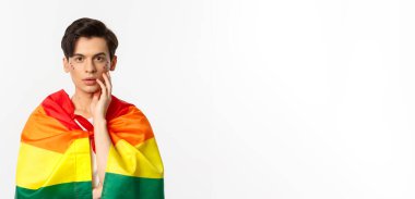 lgbt gurur bayrağı ve parıltısı takan genç, güzel eşcinsel bir adam. Yüzüne dokunuyor ve kameraya şehvetli bakışlar atıyor. Beyaz arka plan..