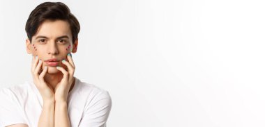 Millet, lgbtq ve güzellik konsepti. Yüzüne mavi ojeli parmaklarıyla dokunan, beyaz arka planda duran güzel eşcinsel adamın yakın çekimi..