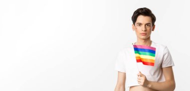 Gurur ve lgbtq kavramı. Gökkuşağı bayrağı tutan, yüzünde parıltı olan ve beyaz arka planda duran çekici ve kadınsı bir adamın belden yukarı pozu..