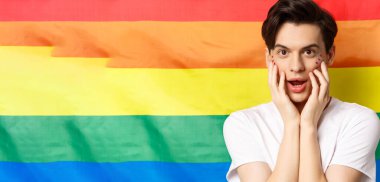 Beyaz tişörtlü, parlak makyajlı, kameraya bakıp eğlenen, yüzüne dokunan, gökkuşağı gurur bayrağına karşı duran güzel esmer gay adamın yakın çekimi..