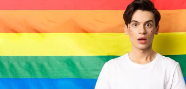 Yüzünde sim olan, kameraya hayret içinde bakan, lgbtq gökkuşağı bayrağının başında dikilen, şaşırmış tuhaf insanın yakın çekimi..