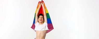Gururlu ve mutlu gay adamın lgbtq gökkuşağı bayrağını dalgalandırması, rahatlamış duygularla gülümsemesi, kot pantolon giymiş, beyaz arka plan.