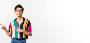 Şüpheli genç adam bakıyor ve şüpheli bir yüz ile sol parmaklarını işaret ediyor, yaz gömleği içinde güvensiz duruyor, beyaz arka plan.