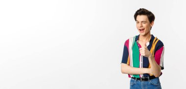 Yakışıklı genç eşcinsel adam baş parmağını kaldırıyor ve göz kırpıyor, seni cesaretlendiriyor, güzel seçimi övüyor, beyaz arka planda duruyor..