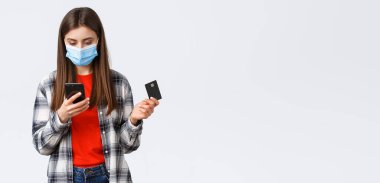 Coronavirus salgını, evden çalışma, online alışveriş ve temassız ödeme konsepti. Tıbbi maskeli bir kız kredi kartı tutuyor, cep telefonu uygulaması kullanarak kıyafet sipariş ediyor..
