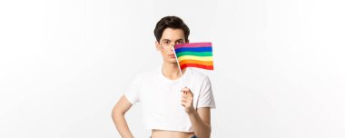 Lgbtq topluluğu. Yüzünde fiske olan çekici ibne, gururlu gökkuşağı bayrağı sallıyor ve kameraya bakıyor, beyaz arka plana karşı mahsul tepesinde duruyor..